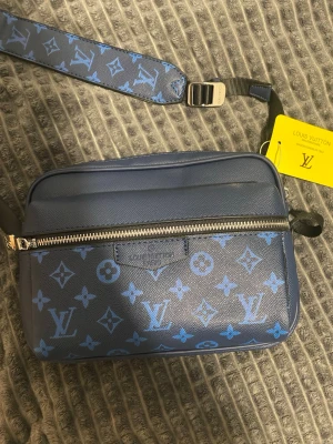 Blå axelväska från L - Snygg blå axelväska från  med klassiskt monogrammönster i ljusblått. Väskan har justerbar axelrem och silverfärgad dragkedja. Tillverkad i tåligt canvasmaterial och har en modern, rektangulär form. Perfekt för dig som vill ha en trendig och praktisk väska.
