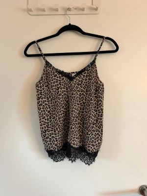 Leopardmönstrat linne med spets - Snyggt loungewear-linne med leopardmönster i beige och svart. Tunna axelband och dekorativ svart spets vid halsringning och nederkant ger en trendig och feminin look. Perfekt för avslappnade dagar hemma. Sparsamt använd