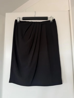 Svart omlottkjol med drapering - Stilren svart kjol med omlottkänsla och snygg drapering framtill. Kjolen har en rak passform och är knälång, perfekt till både sneakers och boots. Tillverkad i ett mjukt tyg som ger ett elegant fall.