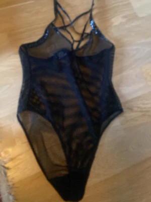 Svart mesh body Killstar - Säljer en svart body från Killstar i meshmaterial med glansiga detaljer och tunna axelband som korsas framtill. Bodyn har ett edgy och alternativt uttryck med transparent tyg och hög skärning vid benen. Perfekt för dig som gillar statement-plagg och unik stil.