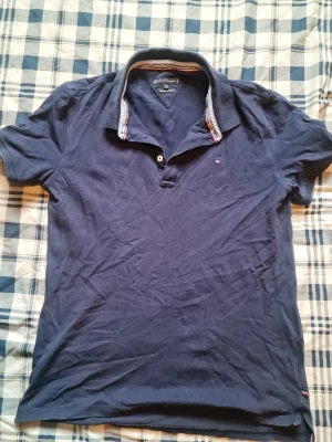 Marinblå pikétröja Tommy Hilfiger - Snygg marinblå pikétröja från Tommy Hilfiger i ekologisk bomull. Klassisk krage med knappslå och diskret logga på bröstet. Perfekt till jeans eller shorts för en avslappnad men stilren look.