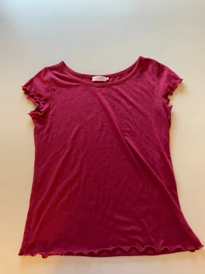 Vinröd t-shirt med volangkant - Säljer en vinröd t-shirt med korta ärmar och volangkant vid ärmslut och nederkant. T-shirten har en enkel, rund halsringning och är gjord i mjuk bomull. Perfekt för en chill och stilren look. Har en liten fläck nertill.