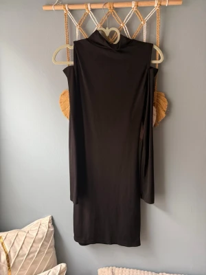 Svart långärmad klänning med drapering - Cool svart klänning med lång ärm och hög krage. Baksidan har en unik draperad detalj med öppningar som ger en edgy look. Klänningen är kort och gjord i ett mjukt syntetmaterial som sitter snyggt på kroppen. Perfekt för dig som gillar statement-plagg.