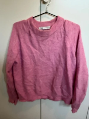 Rosa stickad tröja från Zara - En mjuk och fluffig rosa stickad tröja från Zara med rund halsringning och ribbstickade detaljer vid halsen, ärmslut och nederkant. Perfekt för att skapa en chill och trendig look under kyliga dagar.
