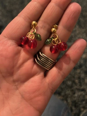Örhängen med körsbär och stål guldfärg - Säljer ett par örhängen med körsbärsdesign. De har röda pärlor som körsbär och små gröna blad, fästa på guldfärgade krokar. Perfekt för dig som gillar lekfulla och färgglada accessoarer.