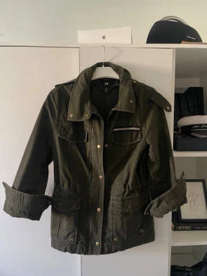 Grön parkajacka från H&M - Säljer en grön parkajacka/safarijacka från H&M med snygga guldknappar och flera fickor. Passar perfekt för våren och kyliga sommarkvällar. Fin Stockholmsstil jacka.