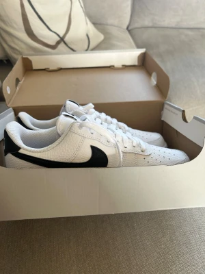 Nike vita sneakers med svart swoosh - Fräscha vita sneakers från Nike med klassisk svart swoosh på sidan. Skorna har lågt skaft, perforerad tå och är tillverkade i läder med textilfoder. Perfekta för en clean och sportig look. Pris ej hugget i sten.