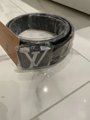 Svart Louis Vuitton bälte med LV-spänne - Snyggt svart bälte från Louis Vuitton med ikoniskt LV-spänne i silverfärg. Bältet är i skinn och levereras med originalförpackning och dustbag. Perfekt accessoar för att lyfta din outfit. Den kommer med kvitto 