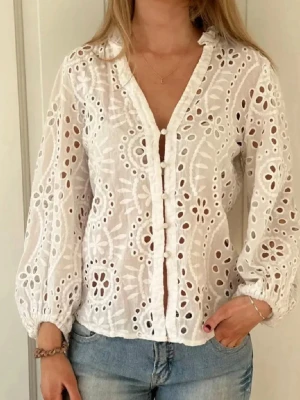 Vit broderad blus med knappar - Superfin vit blus med broderade hålmönster och dekorativa knappar framtill. Blusen har v-ringning, ballongärmar med resår vid ärmslut och är tillverkad i ett lätt, luftigt tyg. Perfekt att styla med jeans för en fräsch look.