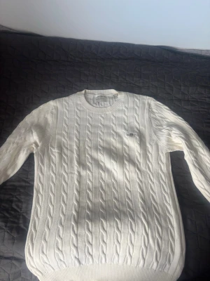 Vit/beige stickat gant tröja  - Stickat vit/beige tröja från GANT med rund halsringning och diskret logga på bröstet. Tröjan har ribbade muddar vid ärmslut och nederkant, och är tillverkad i mjukt material som passar perfekt för lager-på-lager-stil.