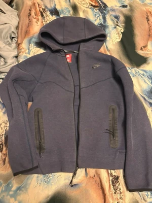 Blå Nike hoodie med dragkedja - Snygg blå hoodie från Nike med hel dragkedja och två fickor med svarta dragkedjor. Hoodien har en sportig look, justerbar huva och diskret Nike-logga på bröstet. Perfekt för chill eller träning. Materialet känns mjukt och stretchigt.