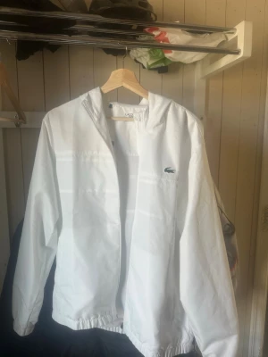 Vit vindjacka från Lacoste med huva - Snygg vit vindjacka från Lacoste med klassisk krokodillogga på bröstet. Jackan har huva, dragkedja framtill och elastiska muddar vid ärmslut och midja. Perfekt för blåsiga dagar och enkel att matcha med olika outfits. Den e sprillans ny, pris kan diskuteras