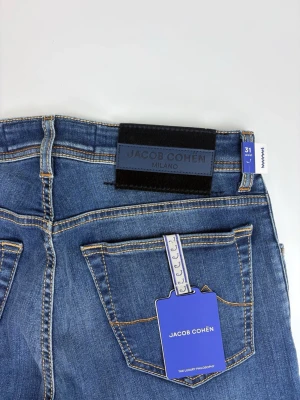 Blå jeans från Jacob Cohën - Snygga blå jeans från Jacob Cohën med klassisk femficksdesign och raka ben. Jeansen har kontrastsömmar och en svart patch med logga bak i midjan. Perfekta för en stilren och avslappnad look. Tillverkade i mjuk denim för skön passform.