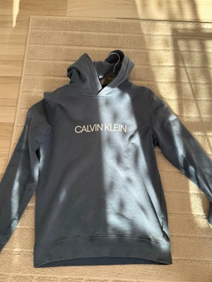 Blå hoodie från Calvin Klein Jeans - Snygg blå hoodie från Calvin Klein Jeans med vit logga tryckt på bröstet. Klassisk modell med huva och ribbade muddar vid ärmslut och nederkant. Perfekt för en avslappnad och trendig stil.