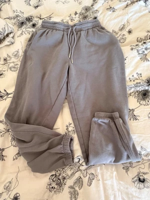 Grå mjukisbyxor med dragsko - Snygga grå mjukisbyxor med resår i midjan och dragsko för perfekt passform. Byxorna har muddar vid bensluten och är tillverkade i ett mjukt och bekvämt material. Perfekta för chill dagar hemma eller när du vill vara extra bekväm.