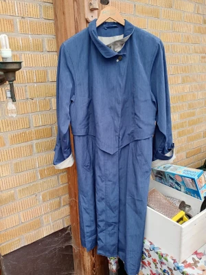 Blå vintage trenchcoat från Goldfix stl 36 - Snygg blå trenchcoat från Goldfix med klassisk krage och knäppning framtill. Lång modell med fickor och uppvikta ärmslut. Ljust innerfoder och retrovibbar, perfekt för dig som gillar tidlös stil och vill sticka ut lite extra.