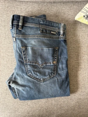 Blå jeans från Diesel - Säljer ett par klassiska blå jeans från Diesel med fem fickor och snygga detaljer som nitar och Diesel Industry-märke på myntfickan. Jeansen har en rak passform och är tillverkade i slitstark denim. Perfekta för en avslappnad och trendig look.