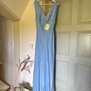 Blå balklänning🩵 - Säljer en så fin helt oanvänd ljusblå långklänning i glansig polyester med djup v-ringning fram och bak. Klänningen har ett smalt knytband i midjan och är ärmlös. Perfekt för balen🩵🩵