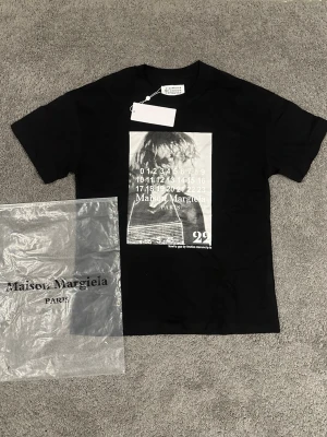 Maison Margiela svart t-shirt - Svart t-shirt från Maison Margiela med tryck framtill i vitt och grått. Klassisk passform med korta ärmar och rund hals. Tillverkad i mjuk bomull och har ikonisk Margiela-etikett i nacken. Perfekt för dig som gillar streetwear och designerplagg.
