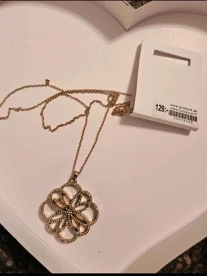 Guldfärgat halsband med blomma - Snyggt guldfärgat halsband från Guldfynd med ett stort blomformat hänge. Hänget har en öppen design med små pärlliknande detaljer längs kanterna som ger extra bling. Perfekt för dig som gillar smycken som sticker ut och vill ha något unikt till din stil.