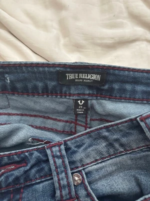 True Religion bootcut jeans blå - Säljer ett par blå bootcut jeans från True Religion med röda kontrastsömmar. Jeansen har låg midja, klassisk femficksdesign och är tillverkade i denim. Perfekta för dig som gillar en avslappnad men trendig look. Pris går att diskuteras 💗