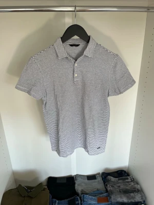 Randig pikétröja från Massimo dutti - Snygg pikétröja från Massimo dutti i vitt och blått med smala ränder. Klassisk krage, korta ärmar och knappslå framtill. Liten logotyp nertill på tröjan. Hör gärna av dig vid frågor!