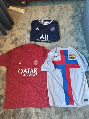PSG och Barcelona fotbollströjor - Tre snygga fotbollströjor: en marinblå PSG tröja med Messi , en röd PSG-tröja med doue på, samt en ljusgrå Barcelona-tröja med röd och blå korsdesign och klubbmärke. Köp individuell har kvitto
