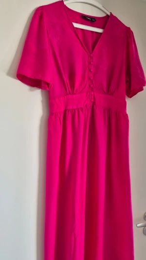 Ceriserosa klänning med knappar märket Pieces från Vila - Superfin ceriserosa klänning från Vila med v-ringning, korta ärmar och dekorativa knappar framtill. Klänningen har markerad midja och är tillverkad i ett mjukt, lätt material som ger ett snyggt fall.
