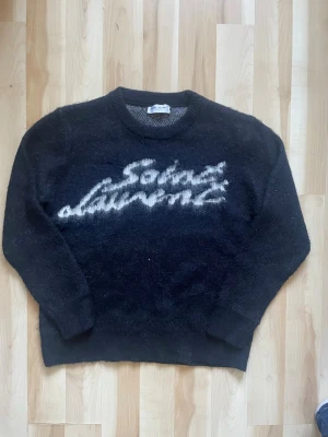 Svart Saint Laurent stickad tröja - Mysig svart stickad tröja från Saint Laurent med vit logotyp i borstigt garn framtill. Tröjan har rund halsringning och långa ärmar. Perfekt för dig som gillar exklusiva och trendiga plagg med streetkänsla.