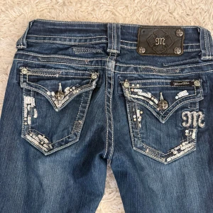Mörkblå Miss Me jeans med paljetter - Snygga mörkblå jeans från Miss Me med bootcut passform. Bakfickorna har detaljerade broderier, paljetter och metallknappar. Jeansen har låg midja och slitningar för en cool look. Perfekta för dig som gillar statement-plagg. Nypris 1499. 