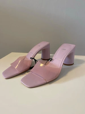 Ljuslila pumps med transparent rem Zara - Säljer ett par ljuslila pumps från Zara med bred transparent rem över foten och fyrkantig tå. Skorna har en chunky klack och öppen häl, vilket ger en modern och trendig look. Materialet är en mix av plast och syntet. Perfekta för dig som vill sticka ut! Oanvända, prislapp kvar och nypris: 399kr.