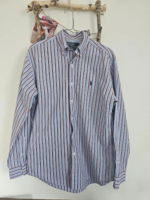 Randig skjorta från Polo Ralph Lauren - Säljer en klassisk långärmad skjorta från Polo Ralph Lauren med blått, vitt och rött randigt mönster. Skjortan har knappar framtill och en broderad logga på bröstet. Perfekt för dig som gillar stilrena plagg med snygga detaljer.