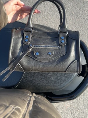 Svart handväska med nitar och dragkedja - Snygg svart handväska i skinnimitation med dubbla handtag, silverfärgade nitar och dragkedjedetaljer framtill. Väskan har en cool, lite rockig look och är perfekt för dig som gillar statement-accessoarer. Praktisk storlek för det viktigaste.