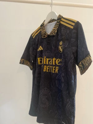 Real Madrid matchtröja svart/guld - Snygg Real Madrid matchtröja från Adidas i svart med guldiga detaljer och mönster. Tröjan har klubbmärke och sponsortryck framtill, samt mönstrad krage och ärmslut i guld. Perfekt för matchdag  eller att bära till vardags.🖤🖤🖤