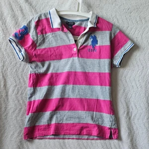 Randig pikétröja från U.S. Polo Assn. - Snygg pikétröja från U.S. Polo Assn. i grått och rosa med breda ränder. Tröjan har korta ärmar, krage med knappar och broderad logga på bröstet. Perfekt för en sportig och avslappnad stil.