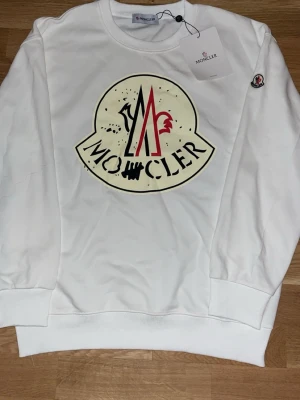 Moncler sweatshirt  - Vit sweatshirt från Moncler med stort tryckt logomotiv i svart, rött och beige på bröstet. Tröjan har rund halsringning, långa ärmar och ribbade muddar. Klassisk och stilren design med Moncler-märke på ärmen. 