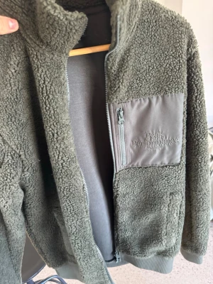 Olivgrön teddyjacka Peak Performance - Mysig olivgrön teddyjacka från Peak Performance med dragkedja framtill och en praktisk bröstficka i matchande tyg. Jackan har hög krage och är tillverkad i fluffigt teddy-material som håller dig varm. Perfekt för lager-på-lager och chill dagar.