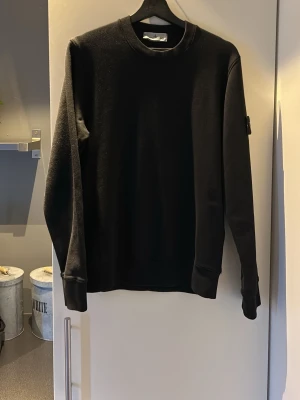 Svart sweatshirt från Stone Island - Svart sweatshirt från Stone Island med rund halsringning och klassisk logotyp-patch på vänster ärm. Tröjan har långa ärmar, ribbade muddar och är tillverkad i mjukt bomullsmaterial. Perfekt för en stilren och avslappnad look.