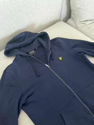 Mörkblå hoodie från Lyle & Scott - Snygg mörkblå hoodie från Lyle & Scott med dragkedja framtill, huva med dragsko och klassisk gul logga på bröstet. Mjuk bomullskänsla och två fickor framtill. Perfekt för en avslappnad och stilren look. Kom med ett rimligt pris så kör vi på det🥰
