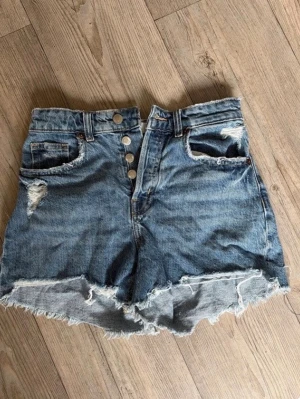 Högmidjade blå jeansshorts med knappar - Snygga jeansshorts i blå denim med slitna detaljer och råa kanter. Shortsen har knäppning framtill med flera silverfärgade knappar och klassiska fickor både fram och bak. Perfekta för sommaren🤗💕