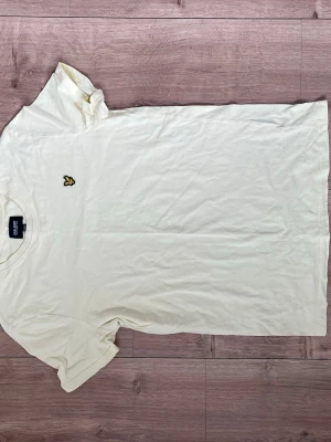 Vit t-shirt från Lyle & Scott - Vit t-shirt från Lyle & Scott med klassisk passform och korta ärmar. T-shirten har en rund halsringning och den ikoniska gula örnloggan broderad på bröstet. Tillverkad i mjuk bomull som är skön att bära hela dagen.