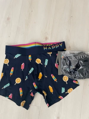 Mörkblå kalsonger med glassmotiv - Säljer ett par mörkblå kalsonger från Happy Socks med färgglada glassar och apelsinskivor som mönster. Midjeresåren har regnbågsfärger och logga. Perfekt för dig som gillar lekfulla och unika underkläder.samt från Kappa som är L