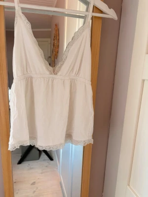 Vit linnetopp med spetsdetaljer - Söt vit linnetopp med smala axelband och v-ringning. Toppen har fina spetsdetaljer längs urringning och nederkant, samt en luftig och avslappnad passform. Perfekt för varma dagar eller som ett lager under en kofta.