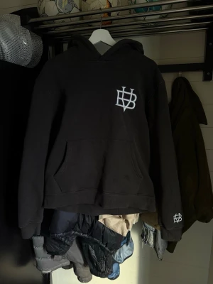 BVALDI hoodie  - Svart hoodie med vit HB-logga på bröstet och ärmen. Klassisk känguruficka framtill och huva. Perfekt för en avslappnad streetwear-look. Mjuk och skön bomullsblandning som passar till det mesta.