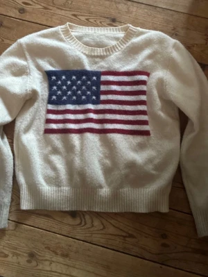 Vit stickad tröja med USA-flagga - Vit stickad tröja med rund halsringning och ribbade muddar. På bröstet finns en stor amerikansk flagga i blått, rött och vitt. Säljer så den inte används längre💕💕
