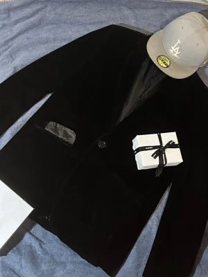 Svart sammetskavaj från Oscar Jacobson - Stilren svart kavaj i sammet från BeeGees med klassisk passform, ett knappfäste och eleganta fickdetaljer i satin. Perfekt för dig som vill ha en dressad look med en lyxig känsla. Fodrad insida för extra komfort. Texturen åvanpå är velvet  och vill vara helt ärlig så disclaimer insidan har lite hål och det syns att den är lite använd men utsidan är jättefin de lovar jag  