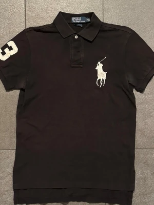 Svart pikétröja från Polo Ralph Lauren - Klassisk svart pikétröja från Polo Ralph Lauren med stor vit broderad logga på bröstet och siffran 3 på ärmen. Tröjan har krage, knappar vid halsen och är kortärmad. Perfekt för en stilren och sportig look. Tröjan är i relativt gott skick men är något solblekt samt mindre fläckar under armarna