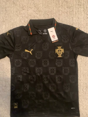 Svart Portugal fotbollströja  limited eusebio edition - Snygg svart fotbollströja från Puma med Portugals landslagsemblem i guld på bröstet. Tröjan har guldiga Puma-loggor, krage med röd och grön detalj samt diskret mönster av Portugals sköld. Tillverkad i lätt och ventilerande material.