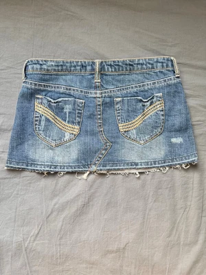 Jeanskjol med slitna detaljer - Kort jeanskjol i blå denim med slitna detaljer och fransig nederkant. Kjolen har klassiska fickor fram och bak, samt dekorativa sömmar och nitar på bakfickorna. Perfekt för en avslappnad och trendig look. Midjemått 76 cm