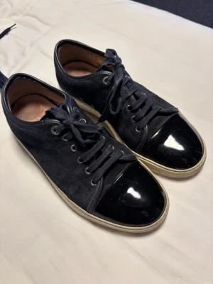 Lanvin mörkblåa - Snygga svarta sneakers från Lanvin med klassisk låg profil. Ovandel i mocka och tåparti i blankt svart lack. Vita sulor och matchande svarta skosnören. Kommer med originalkartong och dustbag. Perfekta för dig som gillar stilrena och exklusiva sneakers. Kom gärna med prisförslag.